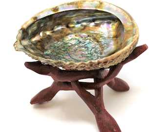 May include: An iridescent abalone shell bowl with a natural, textured exterior and a smooth, multi-colored pearlescent interior, resting on a dark brown, three-legged wooden stand crafted from interconnected, branch-like pieces of wood.