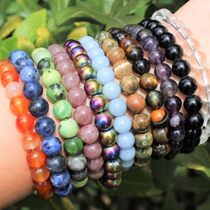May include: A collection of beaded bracelets in various colors, including orange, blue, green, pink, and iridescent. The bracelets are made of polished stones and are displayed on a wrist against a backdrop of green foliage. The beads are approximately 0.3 inches in diameter.