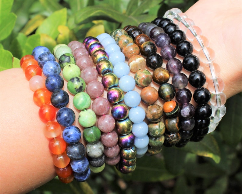 May include: A collection of beaded bracelets in various colors, including orange, blue, green, pink, and iridescent. The bracelets are made of polished stones and are displayed on a wrist against a backdrop of green foliage. The beads are approximately 0.3 inches in diameter.