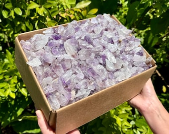 Könnte beinhalten: Eine Kartonbox gefüllt mit einer großen Menge kleiner, roher Amethystkristalle. Die Kristalle sind hellviolett und haben eine durchscheinende Optik.