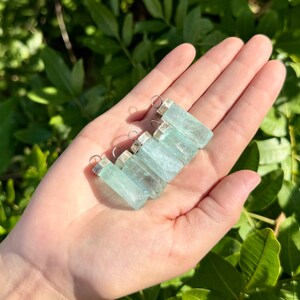May include: A collection of rectangular, light blue aquamarine pendants with silver-colored caps and loops. The pendants are held in an open hand against a backdrop of green foliage, showcasing their translucent quality and natural beauty.