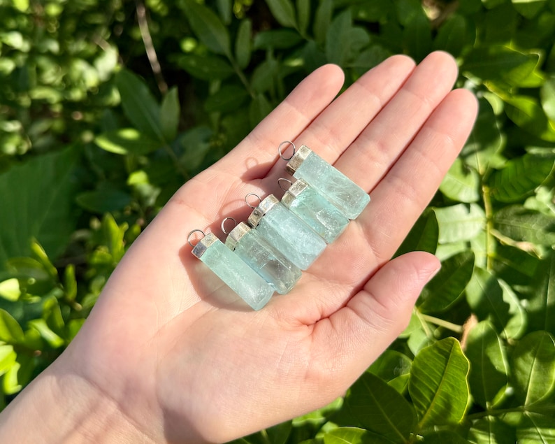 May include: A collection of rectangular, light blue aquamarine pendants with silver-colored caps and loops. The pendants are held in an open hand against a backdrop of green foliage, showcasing their translucent quality and natural beauty.