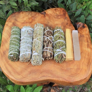 May include: A set of six smudge sticks on a wooden surface. The sticks are tied with white string and include cedar, sage, palo santo, and white sage. There is also a clear quartz crystal and a piece of sandalwood.