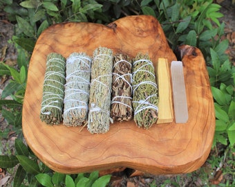 Op de afbeelding: Een set van zes wierookstokjes op een houten oppervlak. De stokjes zijn vastgebonden met wit touw en bevatten ceder, salie, palo santo en witte salie. Er is ook een helder kwartskristal en een stuk sandelhout.