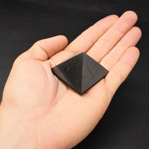 May include: Black obsidian pyramid crystal, a dark, geometric shape, resting in a hand.