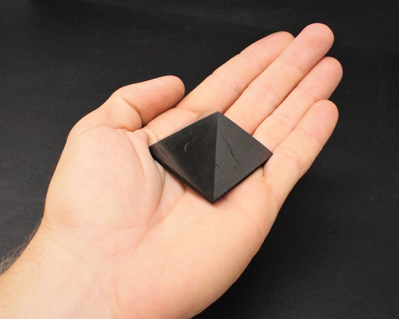 May include: Black obsidian pyramid crystal, a dark, geometric shape, resting in a hand.
