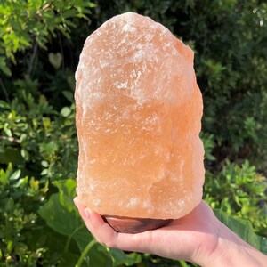 May include: A large, natural Himalayan salt lamp with an orange-pink hue. The lamp is sitting on a wooden base and is being held by a hand.
