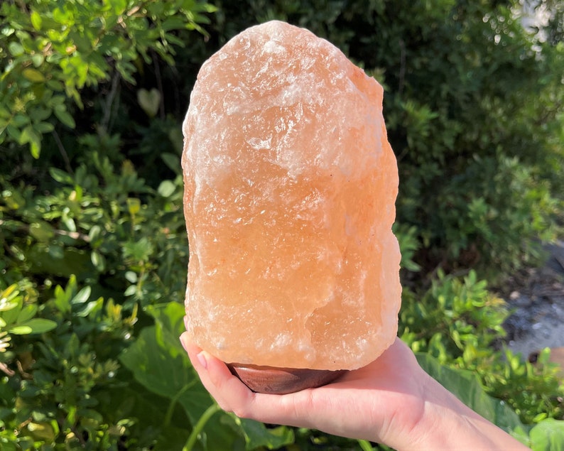 May include: A large, natural Himalayan salt lamp with an orange-pink hue. The lamp is sitting on a wooden base and is being held by a hand.