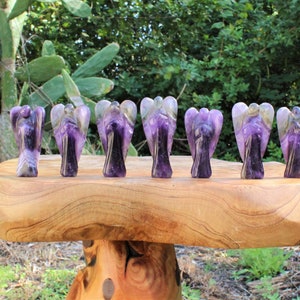 May include: Seven amethyst angel figurines are displayed on a wooden table. The angels have purple and white coloring, with detailed wings and robes. The table is a light brown color and has a natural, organic shape. The background features green foliage.