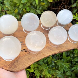 Op de afbeelding: Acht witte selenietkristallen bollen op houten standaards, gerangschikt op een houten plaat. De bollen zijn glad en doorschijnend, met een lichte glans.