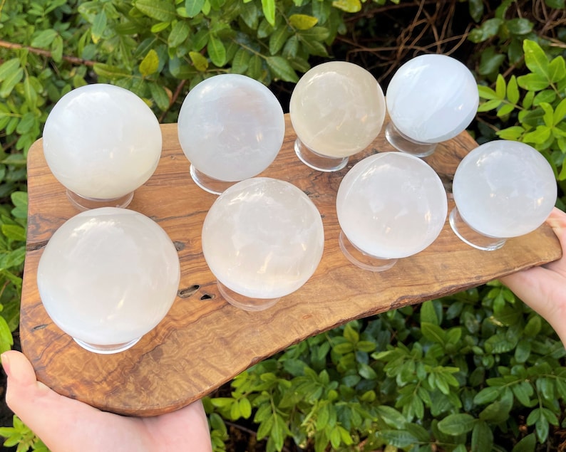 Op de afbeelding: Acht witte selenietkristallen bollen op houten standaards, gerangschikt op een houten plaat. De bollen zijn glad en doorschijnend, met een lichte glans.
