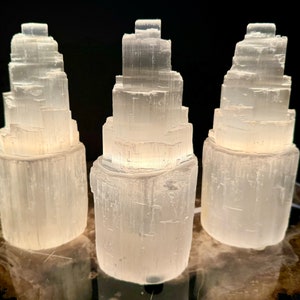 May include: Three white selenite tower lamps with a layered design. The lamps are illuminated from within, casting a soft glow. They are placed on a wooden surface against a dark background, showcasing their natural texture and form.