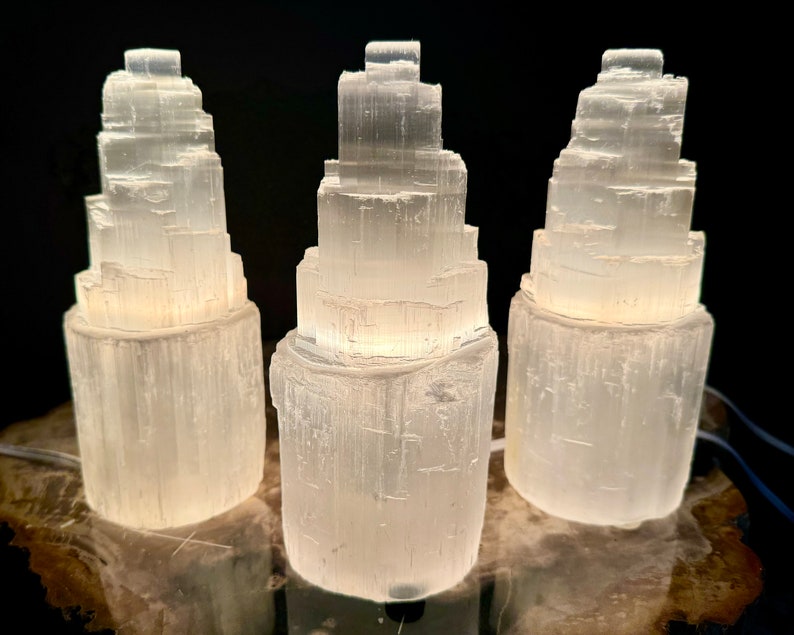 May include: Three white selenite tower lamps with a layered design. The lamps are illuminated from within, casting a soft glow. They are placed on a wooden surface against a dark background, showcasing their natural texture and form.