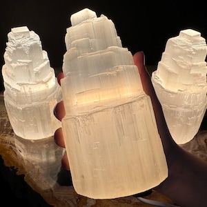 May include: Three illuminated selenite lamps. The lamps are white and have a layered, textured appearance. The lamps are lit from within, creating a soft glow. The lamps are on a wooden surface.