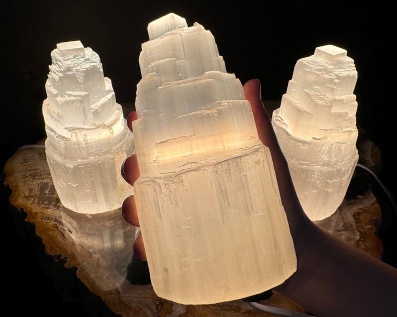 May include: Three illuminated selenite lamps. The lamps are white and have a layered, textured appearance. The lamps are lit from within, creating a soft glow. The lamps are on a wooden surface.