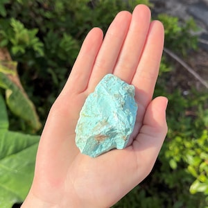 K&ouml;nnte beinhalten: Ein roh geschnittener T&uuml;rkis-Edelstein, der einen lebendigen blau-gr&uuml;nen Farbton mit nat&uuml;rlichen Variationen aufweist. Die unregelm&auml;&szlig;ige Form und Textur des Steins sind sichtbar und unterstreichen seinen rohen, unpolierten Zustand. Der Edelstein wird in einer offenen Hand gehalten.