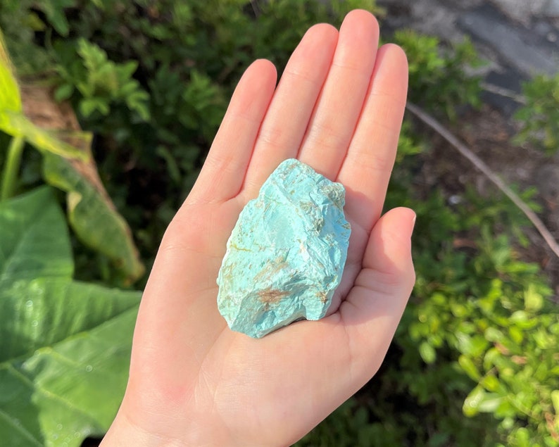 K&ouml;nnte beinhalten: Ein roh geschnittener T&uuml;rkis-Edelstein, der einen lebendigen blau-gr&uuml;nen Farbton mit nat&uuml;rlichen Variationen aufweist. Die unregelm&auml;&szlig;ige Form und Textur des Steins sind sichtbar und unterstreichen seinen rohen, unpolierten Zustand. Der Edelstein wird in einer offenen Hand gehalten.