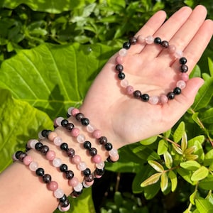 May include: Four bracelets made of pink, black, and white beads. The bracelets are stacked on a person's wrist. The bracelets are made of natural stones.
