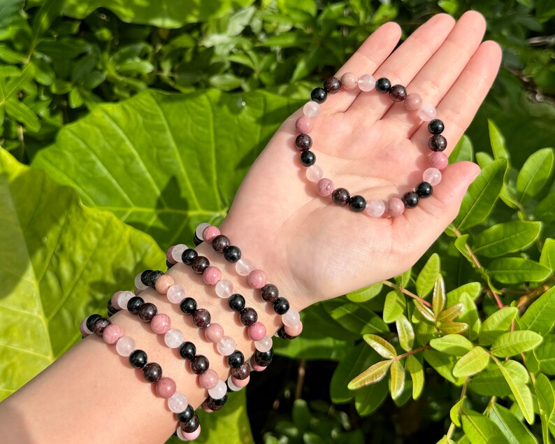 May include: Four bracelets made of pink, black, and white beads. The bracelets are stacked on a person's wrist. The bracelets are made of natural stones.