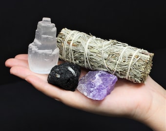 May include: A collection of crystals and a bundle of sage. The crystals include a clear selenite tower, a black obsidian stone, and a purple amethyst.