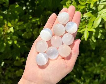 May include: A collection of ten small, smooth, round selenite stones with a translucent, milky white appearance. Each stone features a polished surface that reflects light, showcasing a soft, pearlescent sheen across the cluster of crystals.