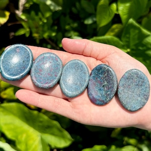 May include: Six smooth, polished, round, blue-green and pink stones with a speckled pattern. The stones are arranged in a row on a hand.