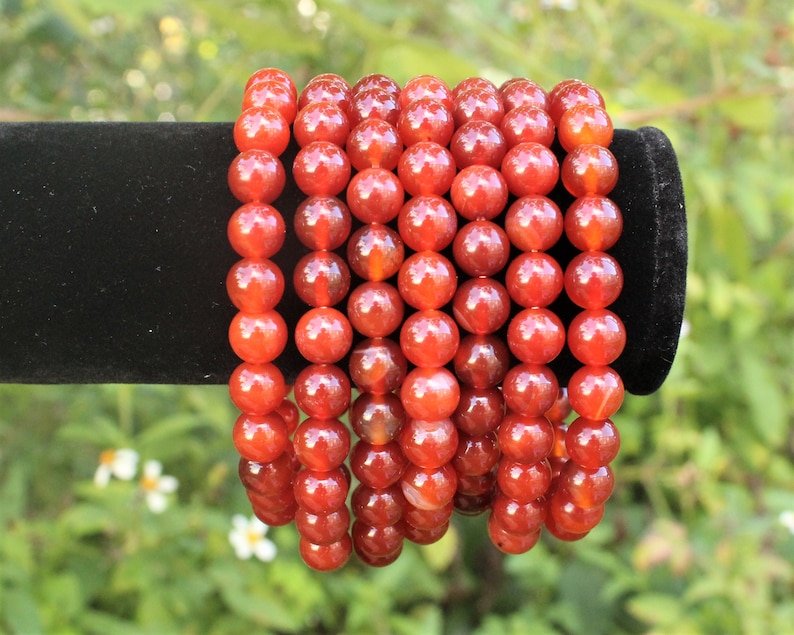May include: Several strands of orange-red gemstone bead bracelets. The beads are round and have a polished, glossy finish. The bracelets are displayed on a black velvet surface against a blurred green background.