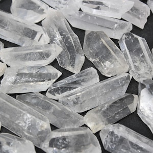 May include: A collection of clear quartz crystal points, varying in size and shape. The crystals are translucent and appear to be naturally formed, with sharp edges and a slightly rough texture. They are scattered on a dark surface.