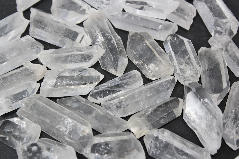 May include: A collection of clear quartz crystal points, varying in size and shape. The crystals are translucent and appear to be naturally formed, with sharp edges and a slightly rough texture. They are scattered on a dark surface.