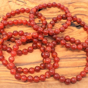 May include: Several strands of red-orange gemstone bead bracelets are displayed on a wooden surface. The beads are round and have a polished, glossy finish. The bracelets are arranged in a circular pattern.