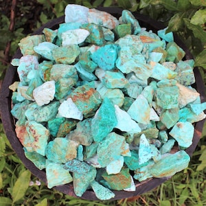 K&ouml;nnte beinhalten: Eine Holzschale gef&uuml;llt mit rohen T&uuml;rkissteinen. Die Steine weisen verschiedene Blau-Gr&uuml;n-T&ouml;ne auf, mit Akzenten von Braun und Wei&szlig;. Die Schale steht vor einem Hintergrund aus gr&uuml;nen Bl&auml;ttern.