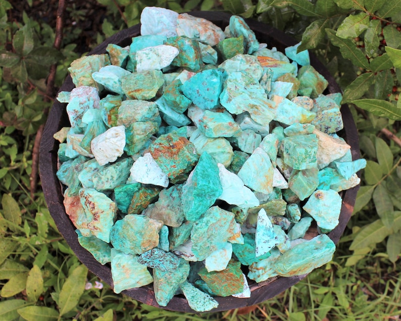 K&ouml;nnte beinhalten: Eine Holzschale gef&uuml;llt mit rohen T&uuml;rkissteinen. Die Steine weisen verschiedene Blau-Gr&uuml;n-T&ouml;ne auf, mit Akzenten von Braun und Wei&szlig;. Die Schale steht vor einem Hintergrund aus gr&uuml;nen Bl&auml;ttern.