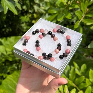 May include: Two beaded bracelets in a small white box. One bracelet is made of black, pink, and white beads. The other bracelet is made of black and pink beads.