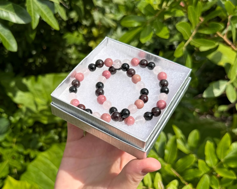 May include: Two beaded bracelets in a small white box. One bracelet is made of black, pink, and white beads. The other bracelet is made of black and pink beads.