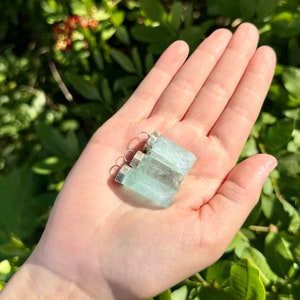 May include: Three aquamarine crystal pendants with silver-colored metal caps and loops. The pendants are a pale blue-green color and are held in a hand against a backdrop of green foliage. The pendants are rectangular in shape.