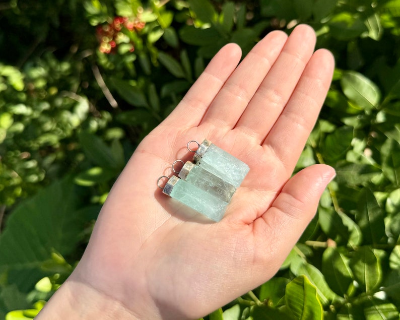 May include: Three aquamarine crystal pendants with silver-colored metal caps and loops. The pendants are a pale blue-green color and are held in a hand against a backdrop of green foliage. The pendants are rectangular in shape.