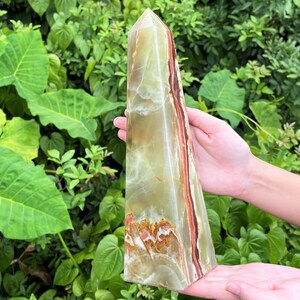 May include: A tall, polished stone obelisk featuring translucent bands of pale green, cream, and deep reddish-brown. The crystal tower has a smooth, faceted surface with natural mineral inclusions and a pointed apex, showcasing a layered colour pattern.