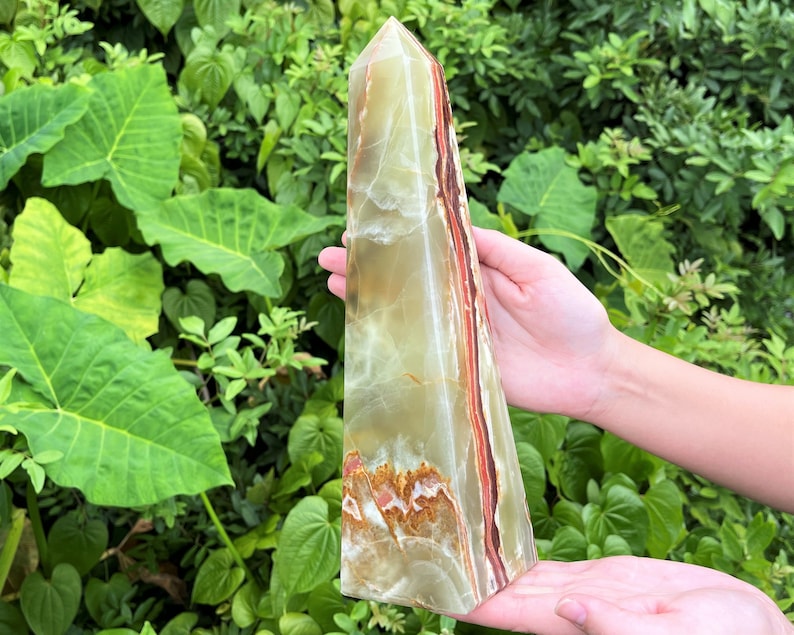 May include: A tall, polished stone obelisk featuring translucent bands of pale green, cream, and deep reddish-brown. The crystal tower has a smooth, faceted surface with natural mineral inclusions and a pointed apex, showcasing a layered colour pattern.