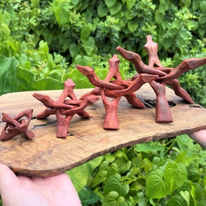 May include: Four reddish-brown carved wooden stands of varying sizes arranged in a row on a natural-edged wooden board. Each stand features a tripod-like structure with interlocking legs and a central cradle designed to support spherical objects.