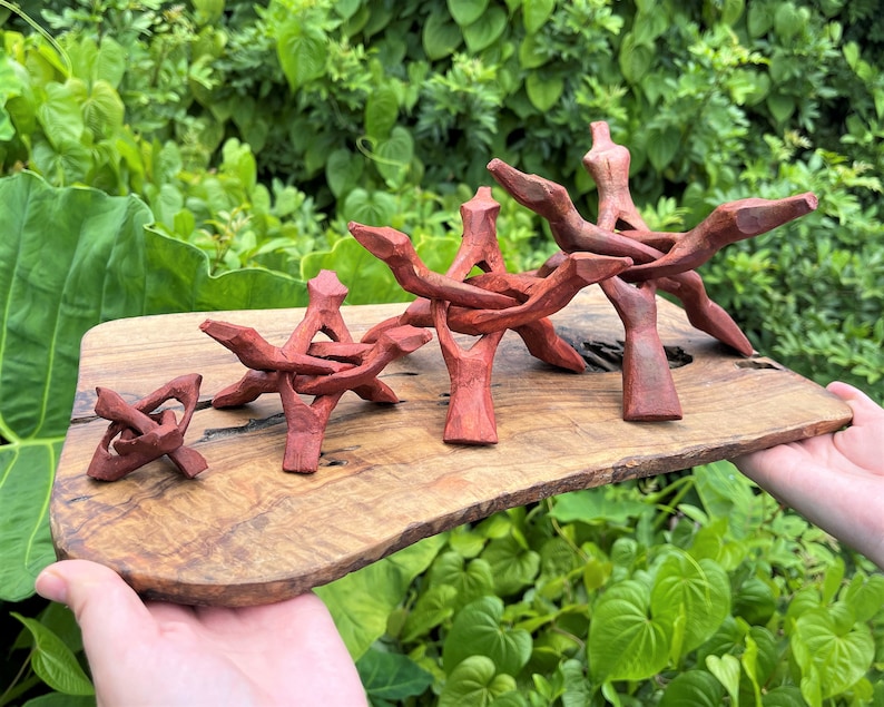 May include: Four reddish-brown carved wooden stands of varying sizes arranged in a row on a natural-edged wooden board. Each stand features a tripod-like structure with interlocking legs and a central cradle designed to support spherical objects.