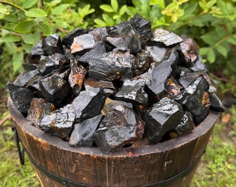 May include: A wooden barrel overflowing with raw, black obsidian stones. The stones have a glossy, wet appearance, with some showing hints of brown and orange. The barrel is made of dark wood and has metal bands.
