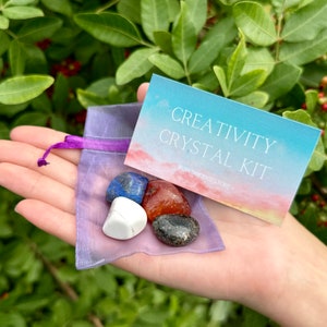 May include: A purple pouch containing a creativity crystal kit with four crystals: blue, red, white, and black. The pouch is in a hand, and a card with the text "Creativity Crystal Kit" is visible.