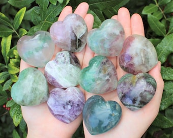 Puede incluir: Una colección de diez cristales de fluorita en forma de corazón en varios tonos de verde, morado y blanco. Los cristales están dispuestos en una mano, con las puntas de los corazones apuntando hacia el espectador.