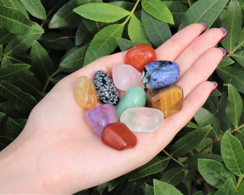 May include: A collection of ten tumbled stones in various colors, including clear, pink, green, blue, brown, orange, and purple. The stones are arranged in a hand, with the palm facing up.
