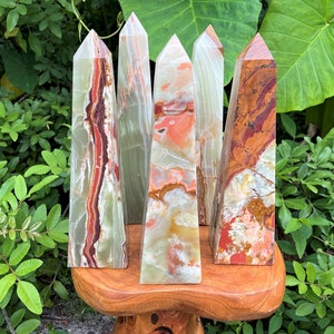 May include: Five polished stone towers with pointed tops, featuring banded patterns in shades of green, orange, red, and white. The towers are arranged in a cluster, displaying smooth, reflective surfaces with natural mineral variations and distinct colour layers.