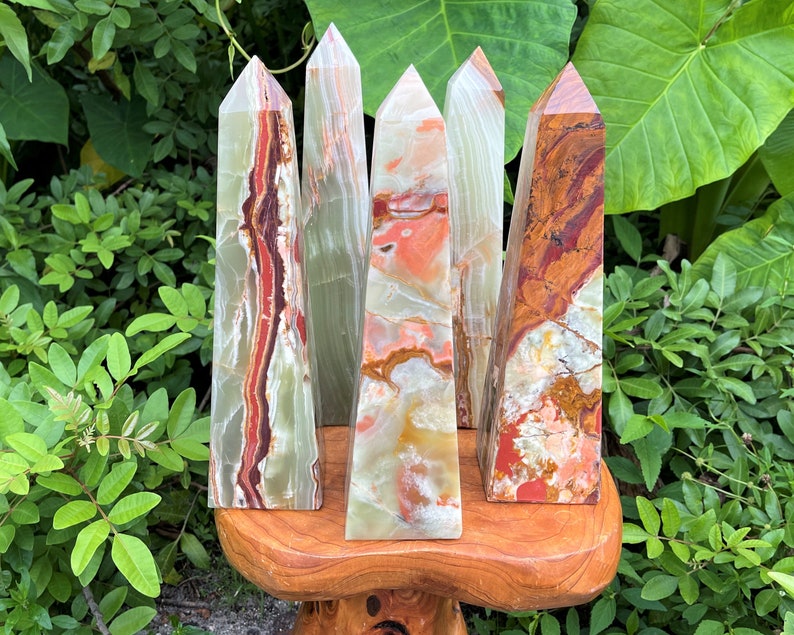 May include: Five polished stone towers with pointed tops, featuring banded patterns in shades of green, orange, red, and white. The towers are arranged in a cluster, displaying smooth, reflective surfaces with natural mineral variations and distinct colour layers.
