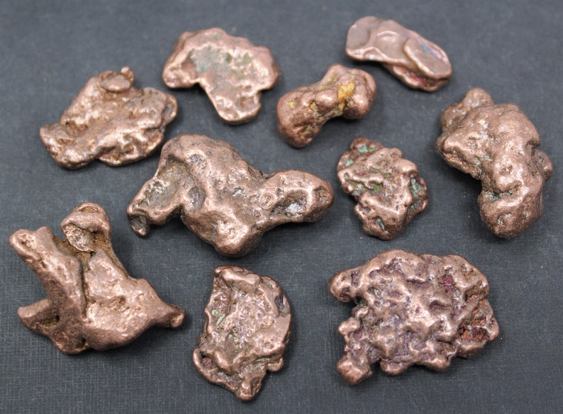 May include: A collection of nine raw copper nuggets with irregular, textured surfaces. The pieces exhibit a metallic reddish-brown color with subtle hints of green oxidation in the crevices, showcasing natural, unrefined shapes and varied sizes.