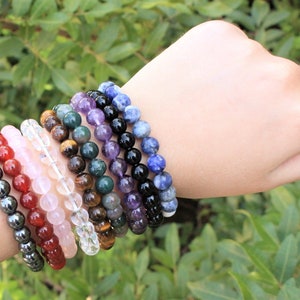 May include: A collection of beaded bracelets in various colors, including red, pink, clear, brown, green, purple, black, and blue. The bracelets are made of gemstones and are displayed on a wrist against a backdrop of green foliage.