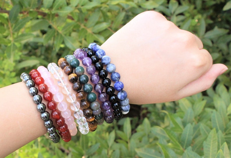 May include: A collection of beaded bracelets in various colors, including red, pink, clear, brown, green, purple, black, and blue. The bracelets are made of gemstones and are displayed on a wrist against a backdrop of green foliage.