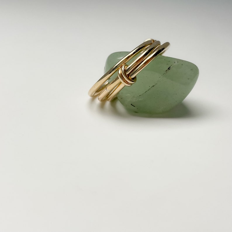 May include: Three gold rings stacked and knotted together, resting on a light green stone.